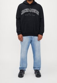 Svart hoodie med framficka och vit logotekst, sportad med ljusblå jeans och svarta sneakers med rundad tå och texturerad sula.