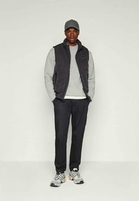 Man wearing gray cap, black sleeveless vest, light gray sweatshirt, black pants, and gray sneakers with orange details standing against a plain background.
