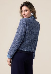 Veste en tweed marine avec une finition texturée, un ourlet effiloché et des poignets boutonnés, associée à une chemise bleue et un pantalon sombre.