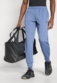 Ανοιχτό μπλε φόρμα με ελαστική μέση και πλάγιες τσέπες, συνδυασμένη με μαύρη τσάντα duffel και μαύρα αθλητικά παπούτσια με λεπτομέρειες από δίχτυ.