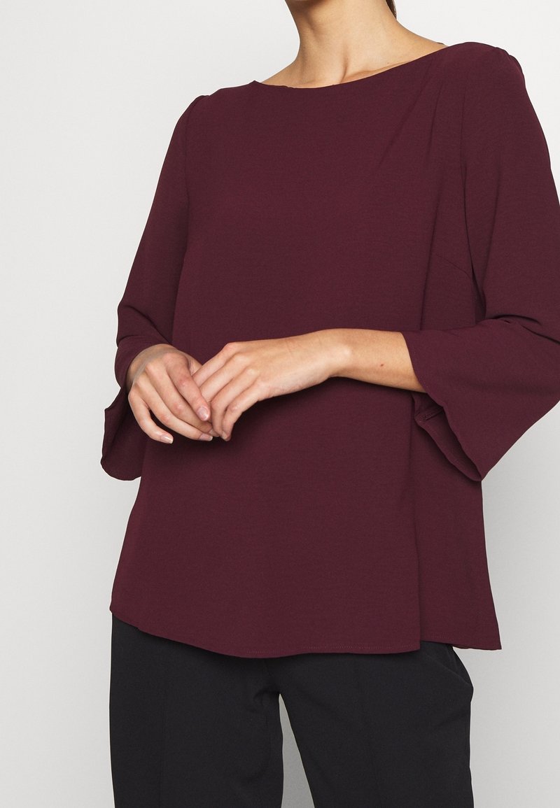 Femme portant un haut à manches longues violet foncé avec des manches larges et un pantalon noir, les mains doucement jointes devant.