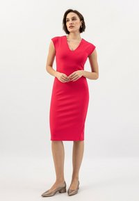Vestito aderente fino al ginocchio di un brillante colore rosa, con scollo a V, maniche corte e una texture liscia. Abbinato a scarpe beige con tacco.
