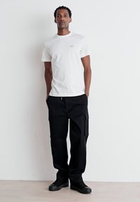 Hvid bomulds t-shirt med et lille logo, parret med sorte cargo bukser med sidelommer, og sorte sneakers på en neutral baggrund.