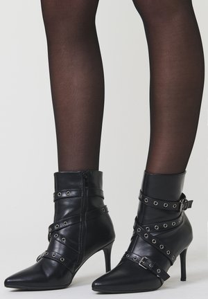 Botines de cuero negro hasta el tobillo con puntas afiladas, tacones de aguja y correas decorativas con ojales metálicos, usados sobre medias negras transparentes.