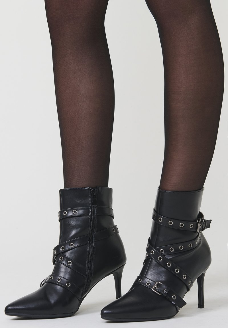 Zwarte leren enkellaarsjes met spitse neuzen, stilettokoppen en decoratieve riemen met metalen oogjes, gedragen over doorschijnende zwarte maillots.
