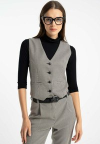 Kobieta w czarnych oprawkach okularów, czarnym golfie, szarym kamizelce w kratę hounds-tooth i pasujących spodniach z czarnym paskiem, stojąca przed białym tłem.