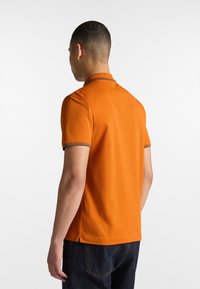 Orange polo skjorte med korte ærmer, der har en krave og kontrasterende trim. Stoffet ser glat ud, med en let tætsiddende silhuet.