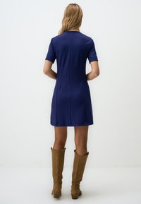 Navyblauw kortjurk met een aansluitend ontwerp en naaddetails, gecombineerd met bruin suède laarzen tot de knie.