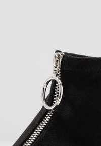 Botte de cheville en suède noir avec un détail de fermeture éclair argentée, dotée d'un tirant circulaire et d'une finition texturée autour de la partie supérieure.