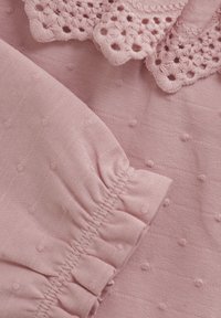 Lys rosa stoff med et teksturert mønster, som har en rynket hekletekstilkant og samlede ermedetaljer, gir dekorative aksenter.