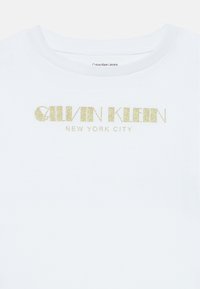 Biała bawełniana koszulka z okrągłym dekoltem i krótkimi rękawami, ozdobiona złotym brokatowym logo z napisem "Calvin Klein New York City".