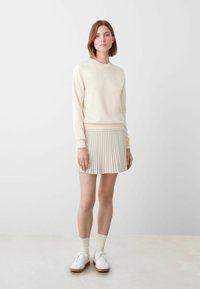 Crème trui met geribde manchetten, gecombineerd met een geplooide beige rok. Witte sneakers en lichte sokken maken de outfit compleet. Neutrale kleurenpalet.