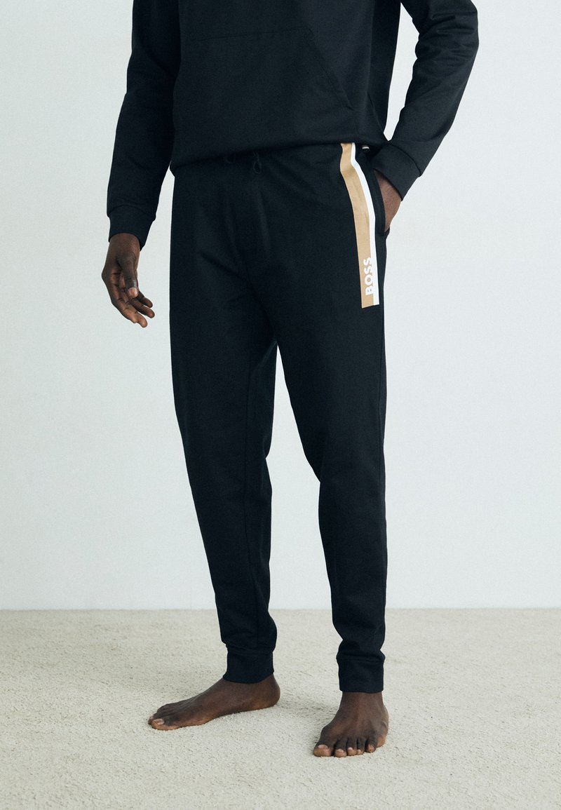 Svart joggebukse med side stripe i gull og hvitt, med elastisk midje og synlig logo på siden. Myk tekstur.
