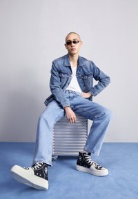 Džínová bunda a světle modré džíny, doplněné černými vysokými teniskami. Model nosí tmavé sluneční brýle, sedí a drží texturovaný kovový předmět.