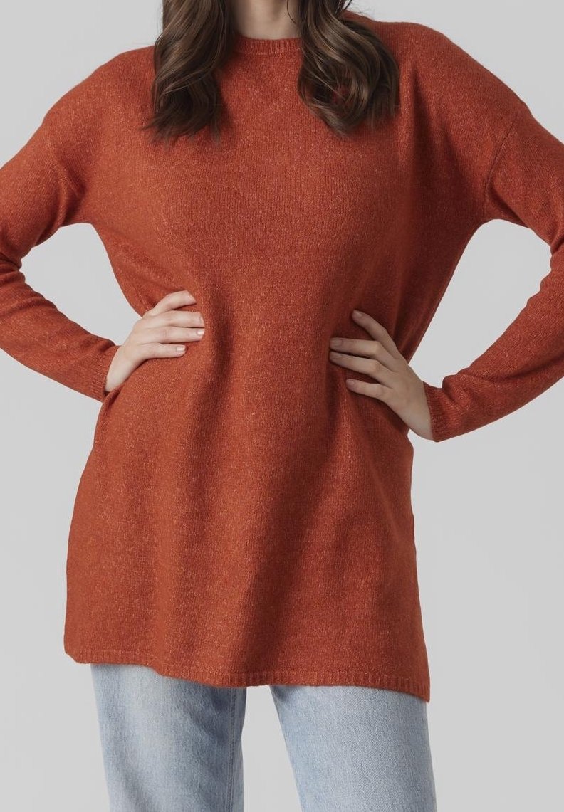 Femme portant un pull à manches longues couleur rouille et un jean bleu clair, debout les mains sur les hanches devant un fond uni.