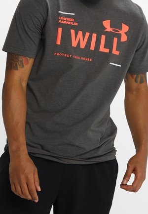 Mand iført mørkegrå Under Armour T-shirt med rød tekst "I WILL" og "PROTECT THIS HOUSE" stående med armene afslappet.