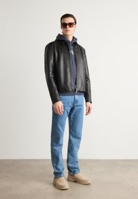 Black leather jacket with a front zipper, gray hoodie featuring a logo, blue jeans, and beige slip-on shoes. Brown sunglasses complete the look.