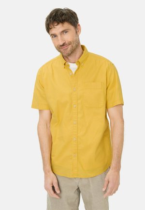 Mand med brunt hår og skæg iført en kortærmet sennepsgul skjorte med knapper og lysebeige bukser, stående mod en hvid baggrund.