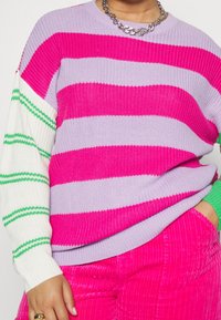 Randig stickad tröja med växlande djärva rosa och ljuslila horisontella ränder, med vita ärmar som är accentuerade av gröna ränder.