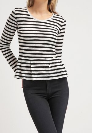 Kobieta w czarno-białym, poziomo paskowanym, długim rękawie bluzce z baskinką, w połączeniu z czarnymi spodniami z wysokim stanem, stojąca na jednolitym tle.