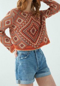 Gestickter Pullover mit Rautenmuster in Orange, Creme und Maroon, kombiniert mit gekrempelten blauen Denim-Shorts.