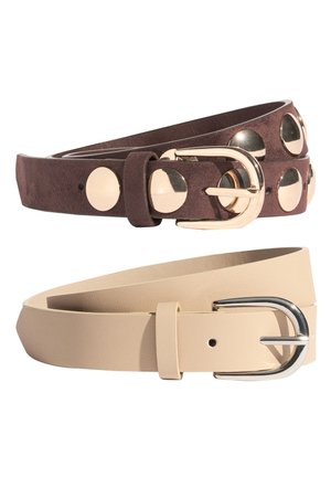 Dva srolovaná kožená páska, jeden tmavě hnědý s velkými zlatými cvoky a zlatou přezkou, jeden béžový se stříbrnou přezkou, na bílém pozadí.