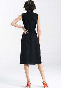 Czarna sukienka bez rękawów z paskiem w talii, długości do kolan, w fasonie A-linii, z teksturowanej tkaniny i subtelnymi plisami z tyłu. Noszona z pomarańczowymi sandałami.