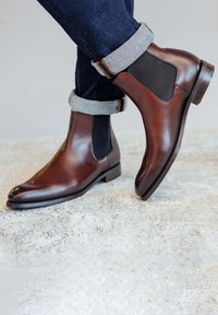 Bottines Chelsea en cuir marron avec des panneaux latéraux élastiques, un design à bout pointu et un petit talon empilé, associées à un jean en denim foncé retroussé.