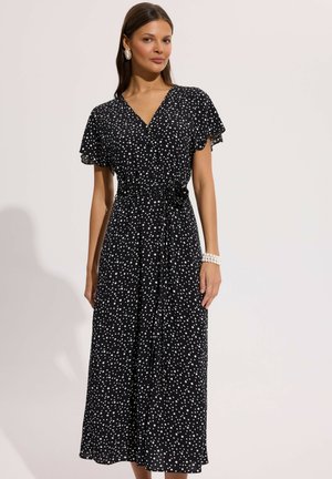 Kobieta w czarnej, krótkorękawowej sukience midi z dekoltem w kształcie litery V, w białe groszki, z podkreśloną talią i perłową bransoletką na neutralnym tle.