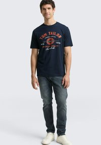 Granatowa bawełniana koszulka z logo "Tom Tailor" w czerwono-białym nadruku. Zestawiona z ciemnymi dżinsami denimowymi i białymi trampkami.