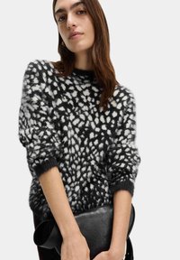 Schwarzer und weißer, flauschiger Strickpullover mit lockerer Passform, der ein gepunktetes Muster und entspannte Ärmel aufweist. Das Modell hält eine schwarze Handtasche.