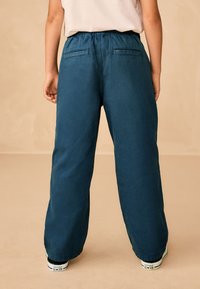 Široké nohavice tmavomodrozelenej farby s elastickým pásom a zadnými vreckami. Nosené s bielo-čiernymi teniskami. Mäkký textilný povrch.