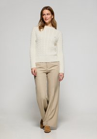 Weißer Kabelstrickpullover mit gerippten Bündchen und Saum, kombiniert mit beigen, weit geschnittenen Hosen. Braune Schuhe sichtbar. Schlichte, klare Linien.