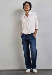 Light pink button-up shirt with a pocket and three-quarter sleeves, paired with dark blue jeans and white sneakers, standing against a gray wall.