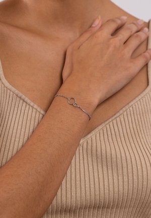 Silbernes Armband mit einem runden Verschluss, das ein zartes Kettendesign aufweist. Am Handgelenk getragen, passend zu einem gerippten, hellbraunen Oberteil.