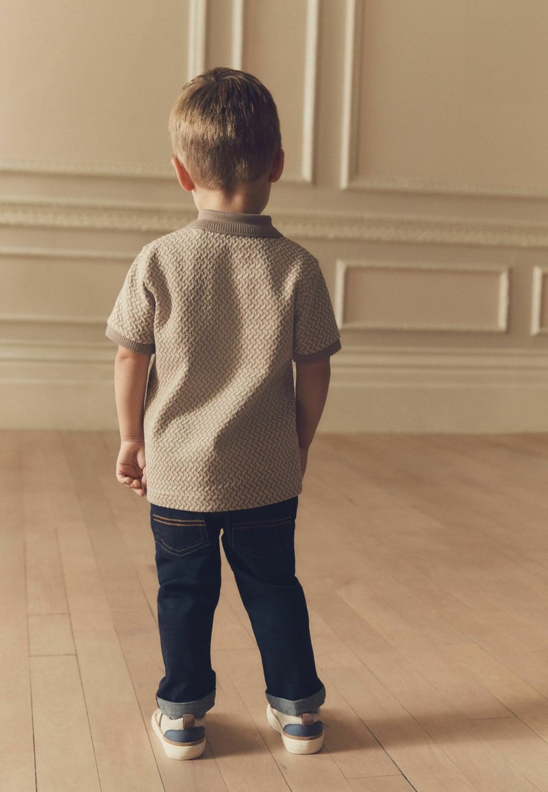 Camisa polo beige texturada para niño con cuello gris, combinada con jeans de mezclilla en azul oscuro. Los zapatos son claros con acentos oscuros. De pie, visto desde la espalda.