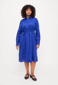 Abito camicia in lino blu con vita cintrata, dettagli con bottoni, maniche lunghe e spacco laterale; indossato con scarpe basse nere.