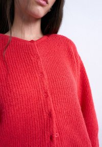 Femme portant un cardigan rouge côtelé à boutons, aux longs cheveux noirs, sur un fond neutre.
