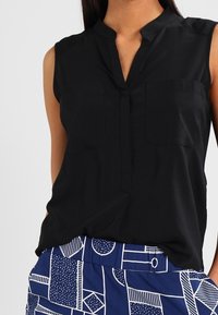 Blusa nera senza maniche con colletto alla cinese, dotata di una patta con bottoni e due tasche sul petto, abbinata a pantaloni blu con motivo.