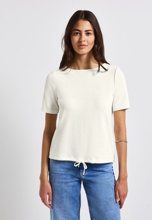 Top bianco in cotone a maniche corte con un motivo testurizzato, scollatura rotonda e orlo con cordino. Abbinato a jeans in denim azzurro chiaro.