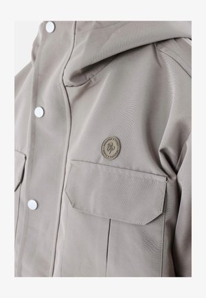 Veste softshell beige avec une fermeture éclair à l'avant, deux poches poitrine et une capuche. Elle est dotée de boutons-pression argentés et d'un petit emblème de marque sur la poitrine.