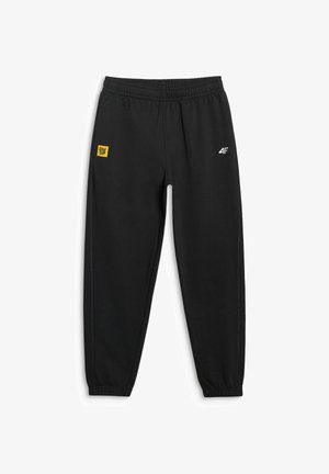 Mężczyzna w czarnych spodniach dresowych marki 4F z elastycznymi mankietami oraz czarnych butach sportowych z czerwonymi akcentami, stojący z rękami w kieszeniach.