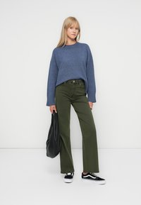 Kék kötött pulóver, khaki széles szárú farmernadrág, fekete sportcipő. A modell egy fekete bőr válltáskát tart. Egyszerű háttér, lágy világítás.