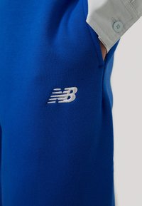 Primo piano di una persona che indossa pantaloni blu brillanti con il logo bianco di New Balance e il polsino della maglia grigio chiaro visibile.