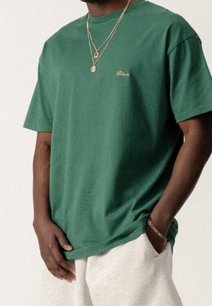 Zielona bawełniana koszulka z krótkimi rękawami, z okrągłym dekoltem i złotym haftowanym logo na lewej piersi. W zestawie z jasnoszarymi szortami.