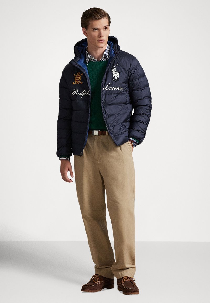Young man wearing navy padded Ralph Lauren jacket, green sweater, striped shirt, beige pants, and brown moccasin shoes, standing against white background.