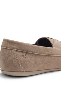 Μπέζ μοκασίνι από σουέντ με λεπτομερείς ραφές και λαστιχένια σόλα, εμφανίζοντας το πλάι και την πίσω φτέρνα με ανάγλυφα αρχικά της μάρκας.