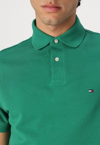 Grøn polo t-shirt med tekstureret stof, knaplukning og et lille rød, hvid og blå logo på venstre brystområde.