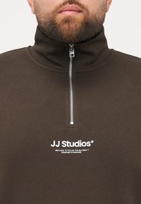Sweatshirt zippé marron avec un col haut, doté d'une fermeture éclair argentée et d'un texte logo blanc sur le devant. Texture lisse, design décontracté.