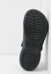 Semelle d'une chaussure Crocs noire montrant un motif de bande de roulement texturé et une indication de taille (W7) sur une surface blanche.